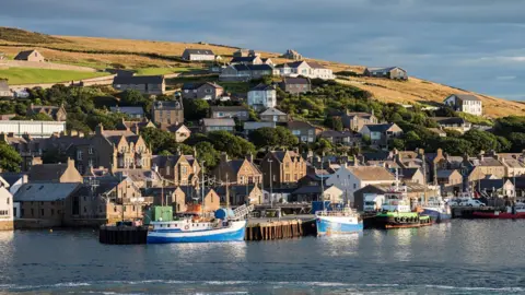 Houses and other buildings in Stromness in Orkney. The properties are on a slope that rises from a harbour. There are fishing boats in the harbour.