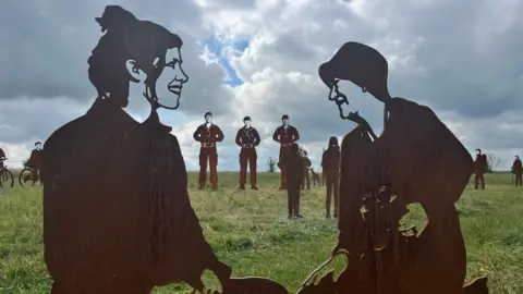 Steel statues of silhouettes on Salisbury Plain. Three soldier statues can be seen in the distance. Statues of two women sat on the grass talking can be seen in the foreground of the image