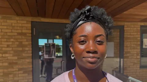 Chris McHugh / BBC Nifemi, a young woman, smiles for the camera. She has silver hooped earrings and black hair held up with a band. A school entrance can be seen behind her.