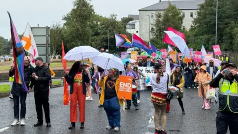 Foyle Pride Lilian Seenoi-Barr at Foyle Pride leading crowd waving pride flags and holding umbrellas in the rain