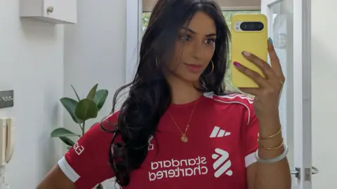 Maz Ullah A woman with long wavy hair smiles as she takes a selfie in a mirror. She is stood in a house and wearing a red football shirt