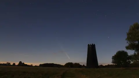 A photo of the grass and trees of Beverley Westwood in silhouette, with the starry skies and comet on display. 