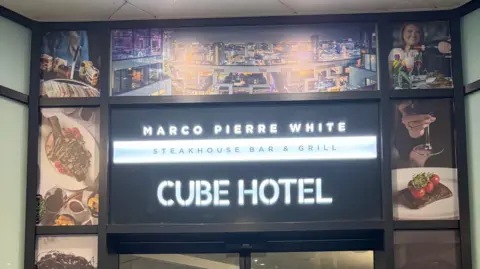 The black and white sign for Marco Pierre White Steakhouse Bar & Grill as well as The Cube Hotel. There are photos of food and drink around it. 
