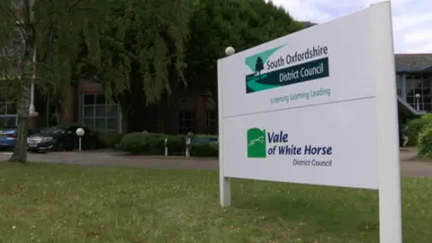 A white sign shows the South Oxfordshire District Council logo and the Vale of White Horse District Council logo. It is situated on a green lawn, in front of a council building. 