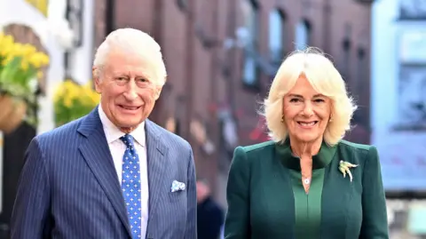 King Charles and Queen Camilla. The king has white hair, wearing blue pin stripe suit, white shirt and blue polka dot tie. The queen has blonde hair, wearing a green coat and gold bird brooch. Both are smiling at the camera