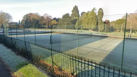 Google A green astro tennis court surrounded by sports chainmail fencing. There are torso high spiked metal railings in the foreground next to a footpath, with the rest of the park, including mature trees in the background.