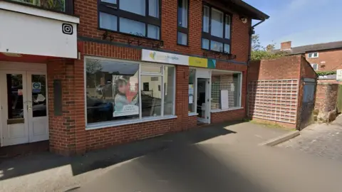 A Severn Hospice site in Leek Street in Wem is in the centre of the photo. A Severn Hospice sign in white, green and yellow is above a ground-floor window in the two-storey building.