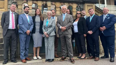 11 people in suits stood infront of grey stone building