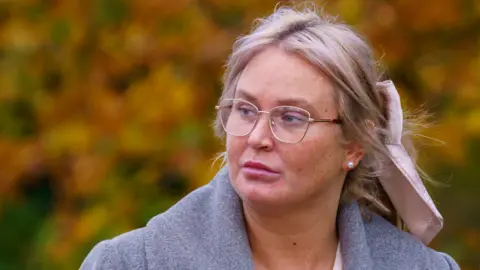 Pacemaker Julie Ann McIlwaine - a woman with blonde hair which is tied back with a light pink bow looks to her right as she is walking. She is wearing silver frame glasses and a grey collared coat. In the background are blurred orange and green trees.