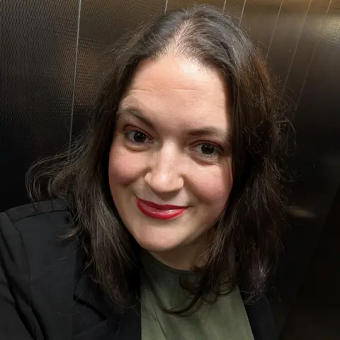 Stacey Panda Selfie dari Stacey Panda di lift. Dia mengenakan atasan hijau di bawah jaket hitam. Dia memiliki bibir merah dan rambut bahu gelap dan menatap kamera.