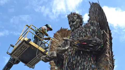 A man on a cherry picker with grey clothing and a helmet facing a large metal sculpture of an angel made from hundreds of knives