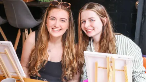 Jess Ward Tiffany Baker Quayle and Caitlin Ball smiling while sitting in front of two small easels. Tiffany is wearing sunglasses and a black tank-top, while Caitlin is wearing a green and white striped shirt. Both have long light brown hair.