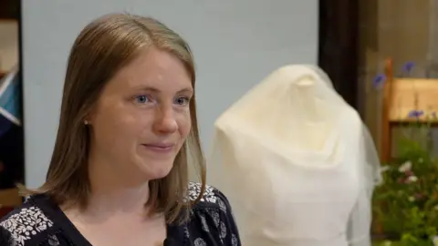 A young woman smiles with a wedding dress in the background.