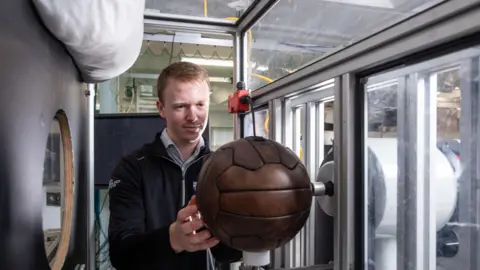 Loughborough University Phillips holding a brown leather ball and placing it into a metal contraption. He has blonde hair and is wearing a black jumper. 