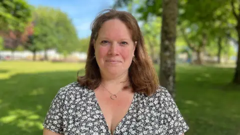 Kate Huntington is looking into the camera while standing in a green park. She has red hair and is wearing a black and white floral top.
