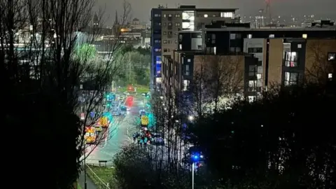 Emergency services lining a street near the large Park Square roundabout. The photo is taken from high up in another apartment building, and some trees block part of the view.