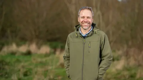 Simon Clarke Simon standing in a field with woodland behind, smiling to camera and wearing a green waterproof jacket and with his hands in his pockets