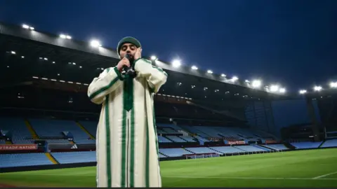 Huzayfa Dawood A man in a robe stands pitch-side at a football stadium.