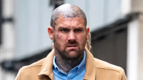 Tony Bartlett walks towards Bristol Crown Court. He is a large man with a shaved head and several tattoos on his neck and head, but not on his face itself. He is wearing a light blue shirt under a light brown jacket.