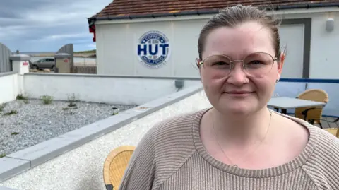 A woman in a beige jumper with glasses and her hair pulled back off her face stands in front of a sign for a cafe