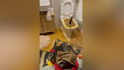 BBC The image shows a toilet overflowing with raw sewage. Brown water covers the bathroom floor. 