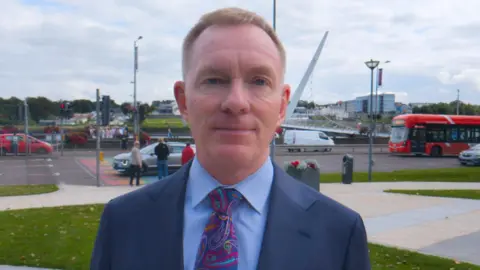  Sir Chris Bryant standing near the Peace Bridge in Londonderry. He is smiling. He's wearing a navy suit with a blue shirt with a multicolouerd tie.