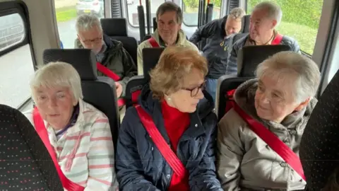A group of older people on board a bus, wearing pink seatbelts.