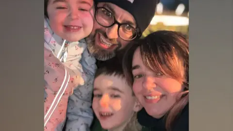 Cillian Kirwan and his wife Louise and two children. They are all grinning at the camera. Cillian is wearing a black beanie, black glasses and his daughter is wearing a white floral coat, his son is grinning next to his mum who has long brown hair. 