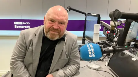 Bill Revans, leader of Somerset Council sitting in the BBC Radio Somerset radio studio looking at the camera.