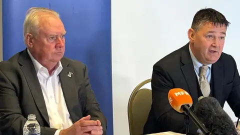 Alan Savage, left, is wearing a grey suit jacket over a white shirt. He is sitting next to joint administrator James Stephen.