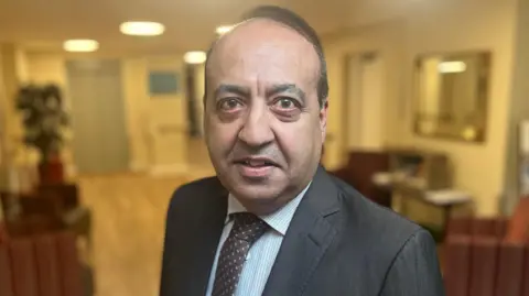 BBC Zuffar Haq wearing a dark grey suit, white and blue striped shirt and dark tie.