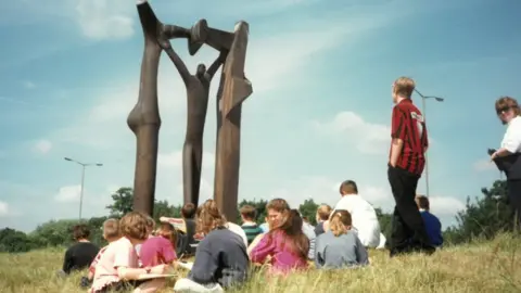 Nene Park Trust A group of children sat on the grass while on a school trip to see the Peterborough Arch sculpture in 1998. The sky is blue with some clouds and the grass is long and looks quite dry. The sculpture stands tall in front of the children and shows an abstract wooden man with his arms up, touching the top of a wooden arch.