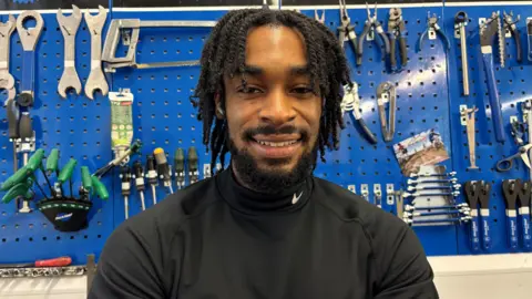 Trevelyan Shonubi in a black jumper. He smiles and stands in front of a blue wall full of bike repair tools.