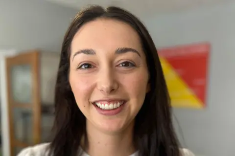 A woman with dark hair and brown eyes wearing a white top smiles into the camera