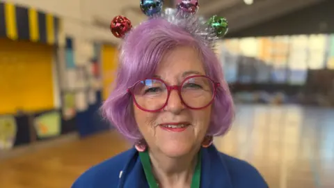 A woman with glasses and purple hair is looking at the camera. She is wearing a blue top and the background is blurred.