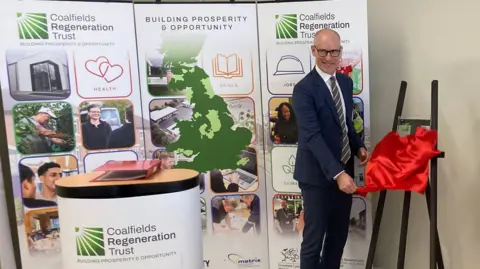 A man wearing a suit stands to the right of the frame. He is wearing a blue suit and a grey striped tie. He is bald and has glasses. He is unveiling a plaque on a stand, covered with a red a sheet. Behind him is an information board about services offered by the Coalfields Regeneration Trust.
