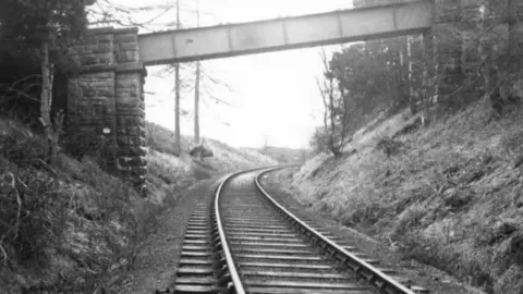 John Mallon/North East Railway Association A black-and-white image of a railway line with a narrow metal bridge crossing it. 