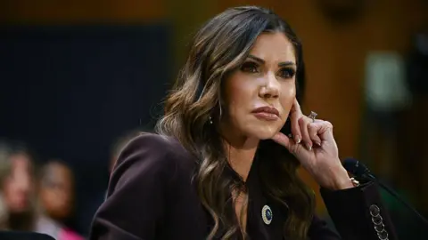 Kristi Noem tilts her head to the right as she props her head up with her left hand. Her hair falls along the side of her face and she is wearing an eggplant coloured blazer with a broach on the left lapel. 