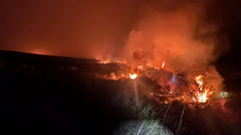 Devon & Somerset Fire & Rescue Service A view of a wildfire at night on Dartmoor. Burning gorse is giving off several sets of flames and smoke.
