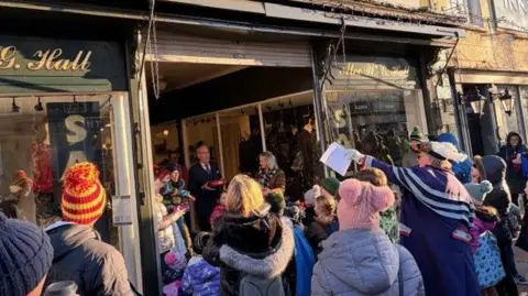 A crowd of children dressed in cold-weather clothing wait outside an old-fashioned shop doorway waiting for a man to drop sweets for them to pick up. Parents wait with them and a councillor in purple regalia with a sheet of paper points towards the man with the sweets.