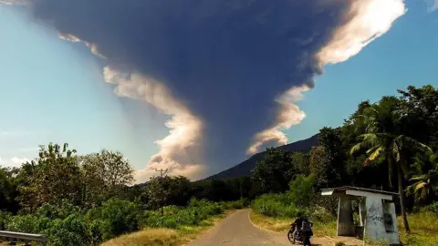 Mount Lewotobi Laki-laki erupts, spewing a huge ash column into the air, as seen from Nangahale village in Sikka, East Nusa Tenggara. Two people on a motorbike are seen in the foreground, along a road leading to the mountain.