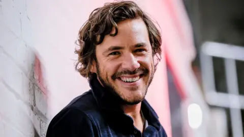 Head and shoulders of Jack Savoretti grinning at the camera while leaning on a brick wall that's painted white. He has medium-length brown wavy hair nd a short beard