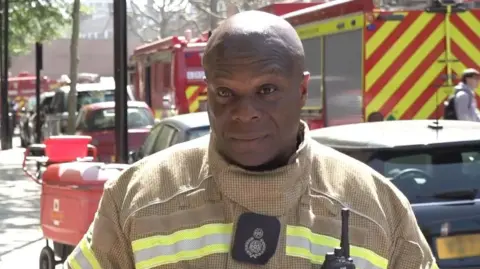 A fire officer in his kit giving an interview to the media at the scene of a fire. There are fire trucks parked in the background.