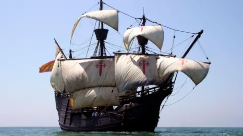 The Nao Victoria Foundation. A replica of the Nao Victoria. It is a dark brown wooden ship. There are six main sails on the boat. Three of them have red crosses on. The ship is in the sea. The sea is calm and blue. The sky is blue and cloudless.