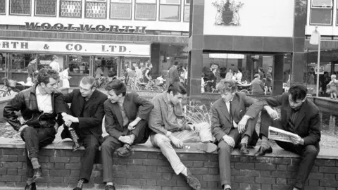 Terry Fincher/Mirrorpix/Getty Images Six young men are sitting on a wall in an old black and white image. One is reading a newspaper and there are shops behind them.