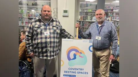 Two men in checked shirts stand either side of a poster on an easel which reads 'Coventry Resting Spaces'