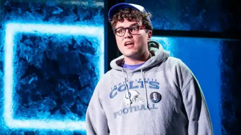 Marc Brenner Tom Dickerson has brown curly hair and glasses. He is wearing a blue and white baseball cap and a grey hoodie with Indianapolis Colts Football written on it. He is singing against a glass effect background that is different shades of blue.