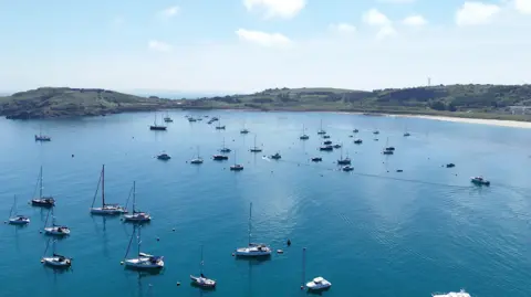 Image shows yachts on calm blue waters with golden sand and green coastline of the island of Alderney.