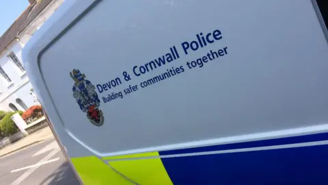 Side view of a Devon & Cornwall Police vehicle featuring the text 'Devon & Cornwall Police – Building safer communities together' alongside a police crest. A yellow and blue stripe runs along the bottom with the message 'For a non-emergency call 101.