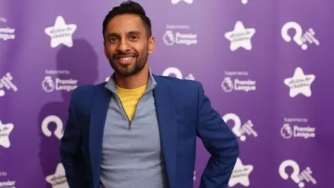 PA Bobby Seagull, a man with short black hair, wearing a yellow t-shirt, with a grey half-zipped jumper and a blue blazer, smiles for the camera while standing in front of a purple background.